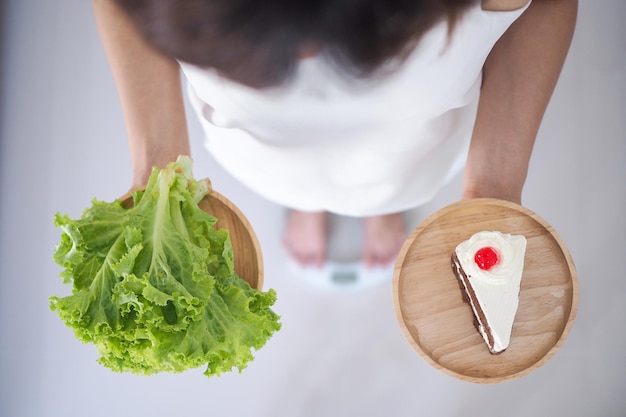 Mujer sosteniendo platos y pasteles de verduras pensando en elegir comer lo que sea más beneficioso para el organismo. Concepto de pérdida de peso, comer cosas que lo hacen saludable.