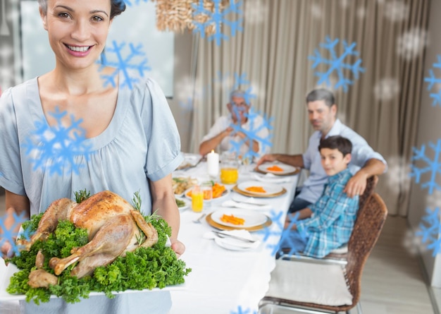 Mujer sosteniendo pollo asado con familia en la mesa de comedor contra copos de nieve