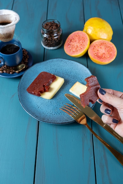 Mujer sosteniendo una rebanada de dulce de guayaba con un bocado junto a frijoles con queso y una taza de café vertical