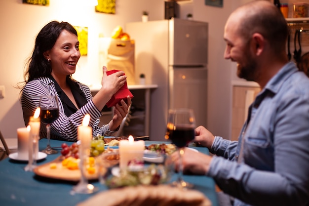 Mujer sosteniendo el regalo de aniversario del marido durante la cena. Feliz pareja alegre cenando juntos en casa, disfrutando de la comida celebrando su aniversario.