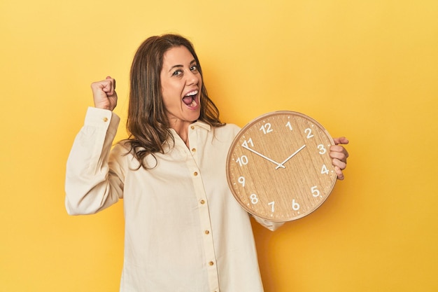 Foto mujer sosteniendo el reloj sobre fondo amarillo levantando el puño después de un concepto ganador de victoria
