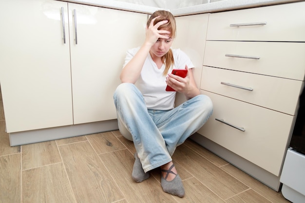 Mujer sosteniendo su cabeza con un teléfono inteligente sentado en el suelo en la cocina problemas depresión