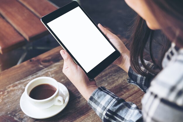 una mujer sosteniendo tablet pc negro con pantalla en blanco blanco con taza de café