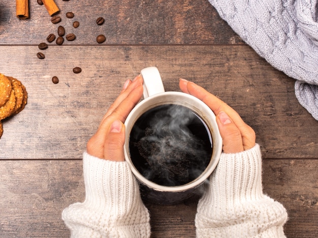 Mujer sosteniendo una taza de café caliente en la mesa de madera rústica, foto en primer plano de las manos