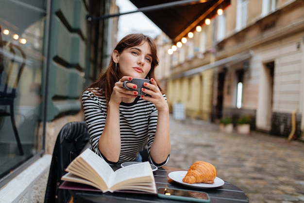 Mujer sosteniendo una taza de café con un libro abierto y un croissant en la acogedora terraza del café