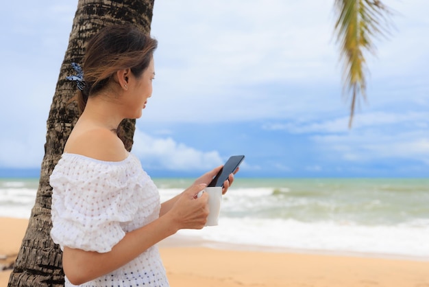 Mujer sosteniendo una taza de café y un teléfono inteligente de pie debajo de las palmeras de coco en la playa del mar y mirando hacia el futuro viendo las olas y el cielo azul con emociones felices y relajantes. Concepto de vida de felicidad y bienestar.