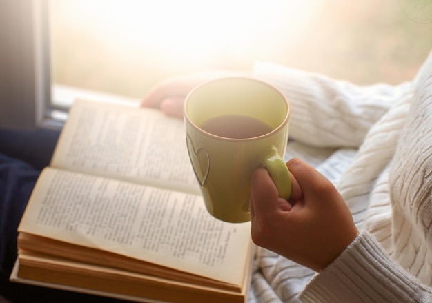 Mujer sosteniendo una taza de té y leyendo un libro al lado de la ventana