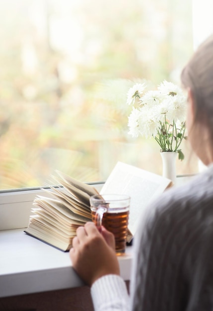 Mujer sosteniendo una taza de té y leyendo un libro al lado de la ventana