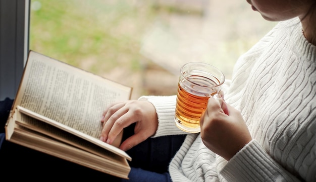 Mujer sosteniendo una taza de té y leyendo un libro al lado de la ventana