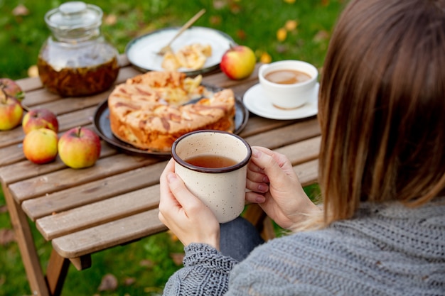 Mujer sosteniendo la taza de té y sentado junto a la mesa con tarta de manzana en el jardín