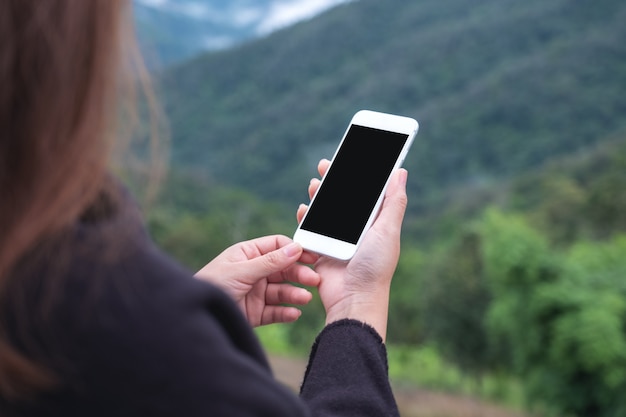 una mujer sosteniendo un teléfono inteligente blanco con pantalla en blanco en el exterior