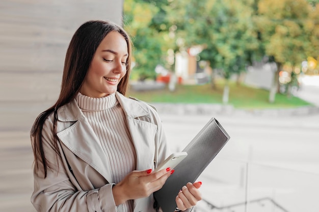 Mujer sosteniendo un teléfono inteligente y una carpeta de un edificio de oficinas