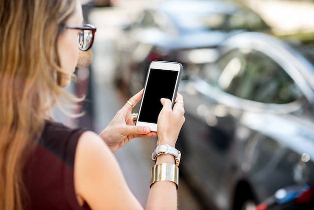 Mujer sosteniendo un teléfono inteligente con pantalla vacía para copiar y pegar con carga de coche eléctrico en el fondo