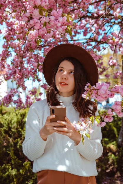 Mujer sosteniendo el teléfono en la mano de pie bajo el floreciente árbol de sakura se acerca la primavera
