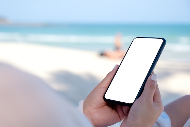 Una mujer sosteniendo un teléfono móvil blanco con pantalla de escritorio en blanco con fondo de cielo azul y mar en la playa