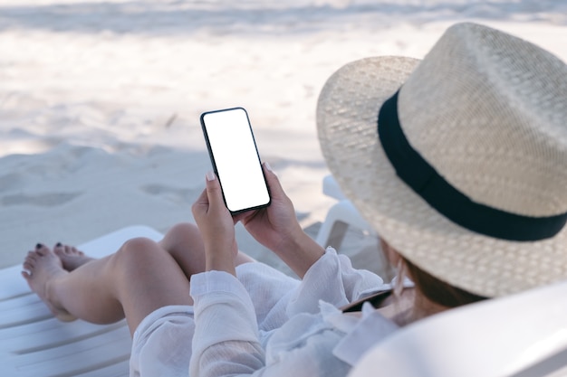 Una mujer sosteniendo un teléfono móvil blanco con pantalla de escritorio en blanco mientras se acuesta en una silla de playa en la playa