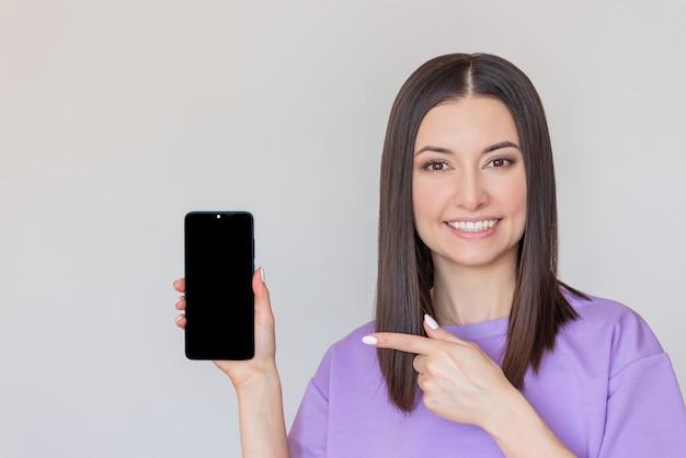 mujer sosteniendo un teléfono móvil en la mano y señalando la pantalla negra y sonriendo Teléfono Mockup