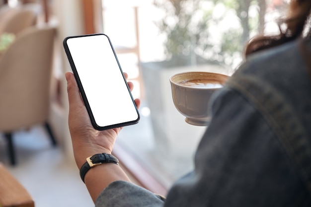 Mujer sosteniendo un teléfono móvil negro con pantalla en blanco mientras bebe café en la cafetería