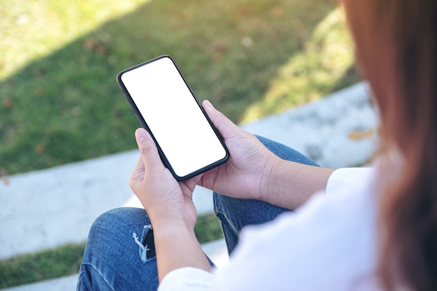 Mujer sosteniendo un teléfono móvil negro con pantalla en blanco mientras está sentado en el parque