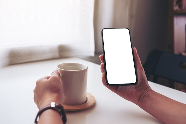 Una mujer sosteniendo un teléfono móvil negro con pantalla de escritorio en blanco mientras bebe café