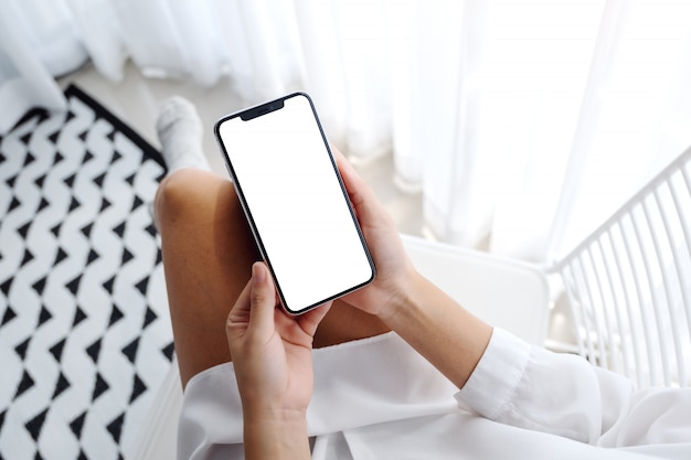 Mujer sosteniendo teléfono móvil con pantalla en blanco de escritorio en blanco mientras está sentado en el dormitorio con sensación de relajación