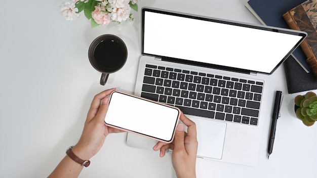 Mujer sosteniendo teléfono móvil con pantalla en blanco sobre la computadora portátil en el escritorio de oficina.