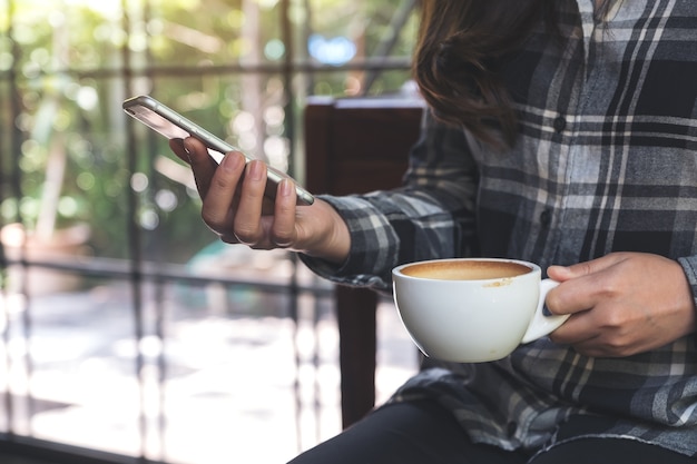 mujer sosteniendo, usando y mirando el teléfono inteligente mientras bebe café