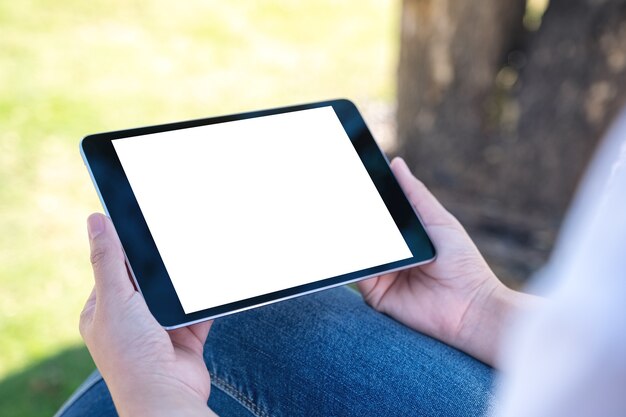 Mujer sosteniendo y usando tablet pc negro con pantalla de escritorio en blanco horizontalmente mientras está sentado en el parque