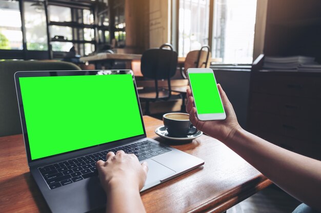 Una mujer sosteniendo y usando el teléfono móvil maqueta con pantalla verde en blanco y una computadora portátil en la mesa de madera en la cafetería