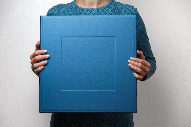 La mujer sostiene un álbum de fotos familiar en una elegante caja cuadrada de diseño. manos femeninas que sostienen la caja cuadrada de la foto para el álbum de boda. Gran caja de regalo azul en manos de mujer de cerca con copia espacio para texto