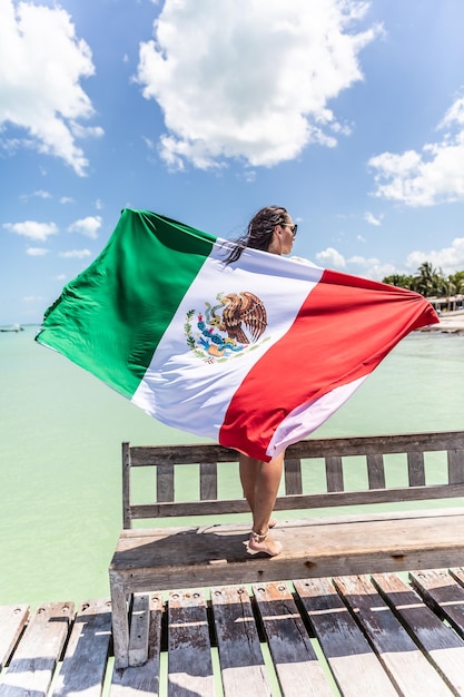 Una mujer sostiene la bandera mexicana a sus espaldas mientras se para en un banco en un muelle al lado del Mar Caribe