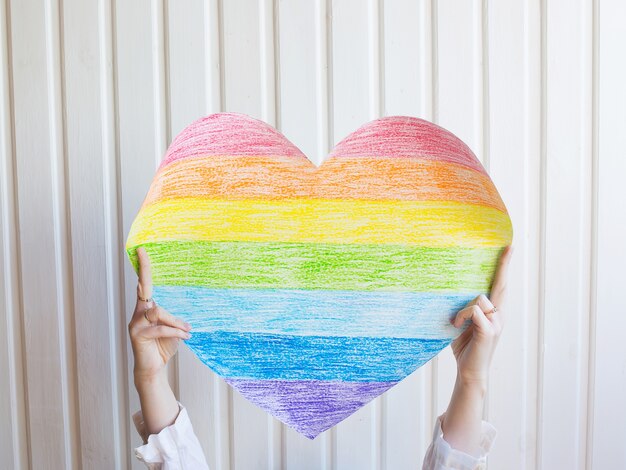 Una mujer sostiene un corazón de arco iris en la pared blanca Símbolo de la comunidad LGBT