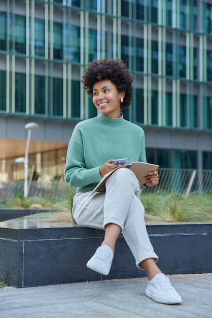 La mujer sostiene el diario y el teléfono inteligente comprueba las notas hace planes para el día sonríe ampliamente vestida con ropa casual plantea contra el edificio de la ciudad bckground