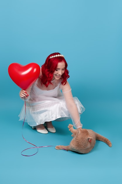 Mujer sostiene un globo volador con forma de corazón, se pone en cuclillas y juega con un gato británico rojo