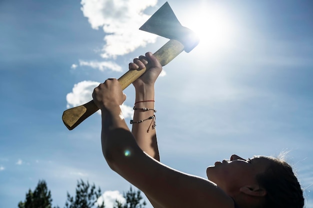 Una mujer sostiene un hacha en sus manos contra el cielo azul, el sol y las copas de los árboles.