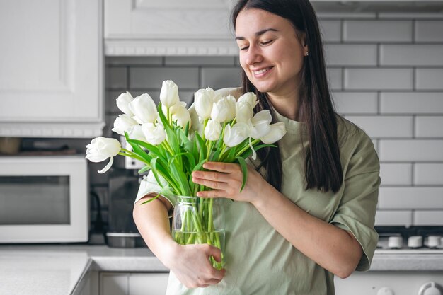 Una mujer sostiene un jarrón con tulipanes blancos en un interior de la cocina