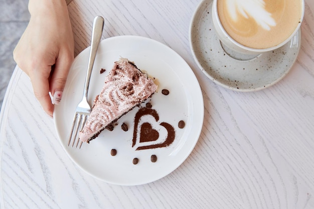 Una mujer sostiene un pastel de chocolate con frutas y una taza de capuchino en una mesa de madera afuera en la terraza del café Desayuno atmosférico Decoraciones de postres en forma de corazón