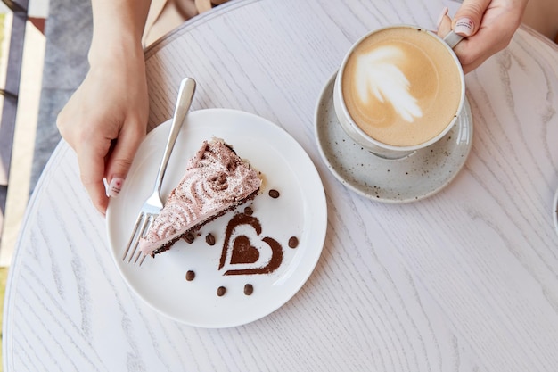 Una mujer sostiene un pastel de chocolate con frutas y una taza de capuchino en una mesa de madera afuera en la terraza del café Tiempo de café atmosférico Tiempo para ti Decoraciones de postres en forma de corazón