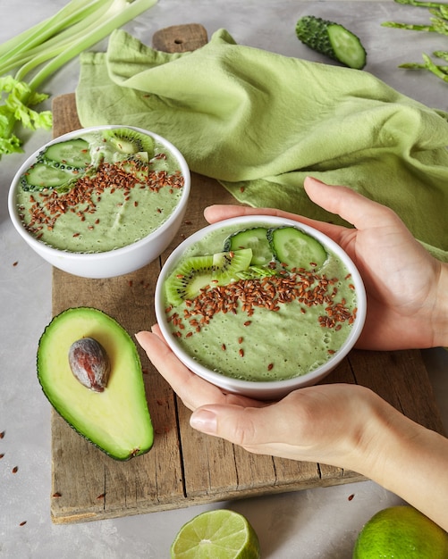 Una mujer sostiene un plato de batidos de apio, aguacate, kiwi y pepino con semillas de lino en el fondo de una tabla de cocina de madera. Un desayuno saludable