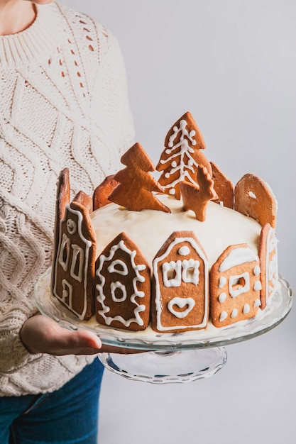 Mujer sostiene tarta casera con fachadas de casa de la ciudad de pan de jengibre, un árbol de Navidad y renos en soporte de vidrio