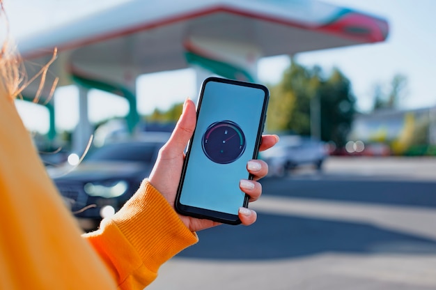 La mujer sostiene un teléfono inteligente con un medidor de combustible digital en la pantalla en el fondo de una gasolinera y un automóvil.