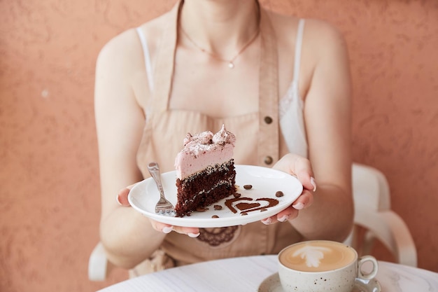 La mujer sostiene un trozo de pastel de chocolate con frutas y una taza de capuchino en una mesa de madera afuera en la terraza del café Tiempo de café atmosférico para ti Decoraciones de postre en forma de corazón