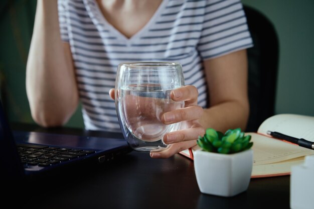 La mujer sostiene un vaso de agua mientras trabaja en el lugar de trabajo de su casa