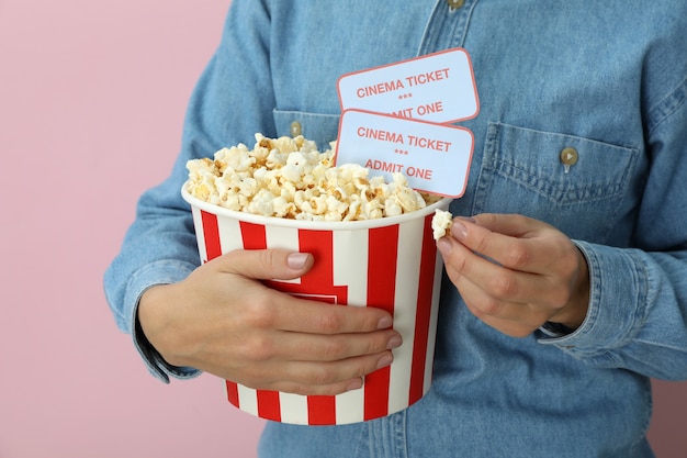 La mujer sostiene un vaso de papel con palomitas de maíz y boletos sobre fondo rosa.