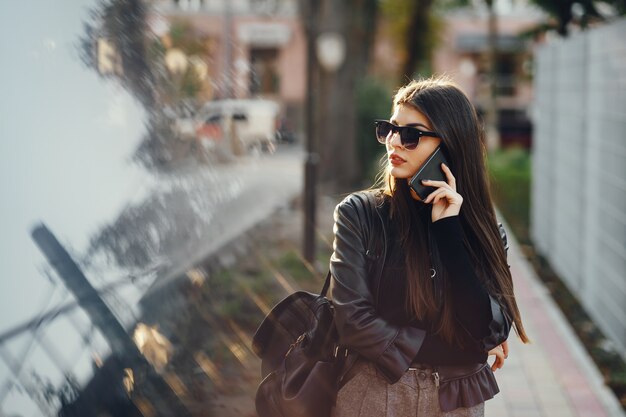 La mujer styligh usa su teléfono celular mientras camina por la ciudad en un día cálido y soleado