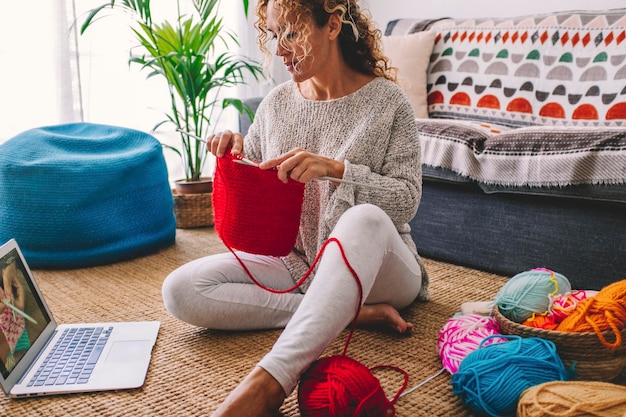 Una mujer en su casa en una actividad de ocio relajada haciendo trabajo de punto con lana roja viendo un video tutorial web en una computadora portátil. Contenido de creador de personas modernas o concepto de clase de lección en línea