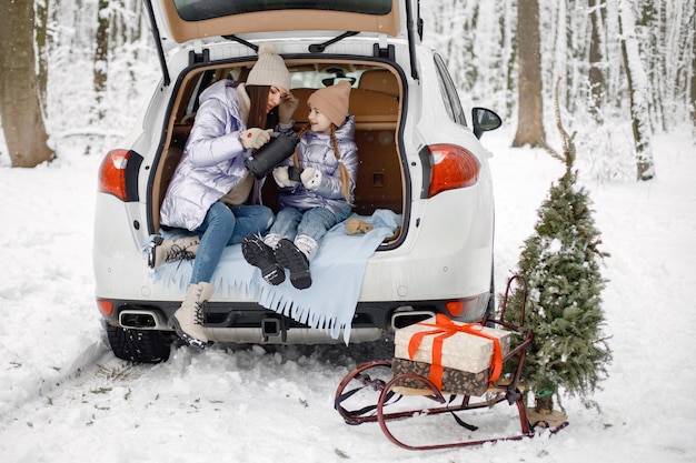 Mujer y su hija sentadas en el maletero del coche abierto en el bosque de invierno