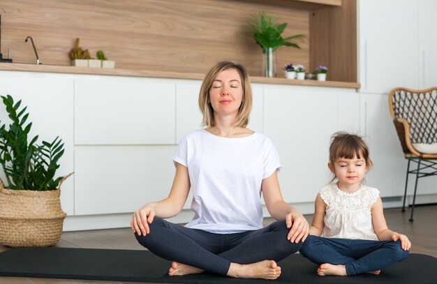 Mujer y su pequeña hija están sentadas en posición de loto con los ojos cerrados mientras hacen yoga juntos en casa