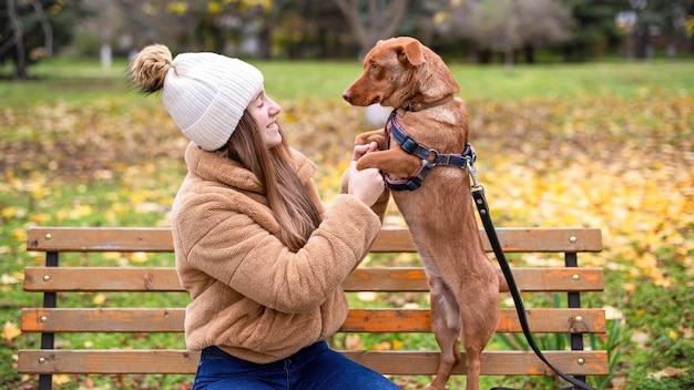 Mujer con su perro en otoño en un parque. Patas de perro en manos de mujer, ella lo mira y sonríe. Banco, césped