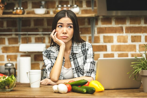 Mujer subrayada asiática hermosa joven con la computadora portátil en cocina. Trabajando a casa. En estres.